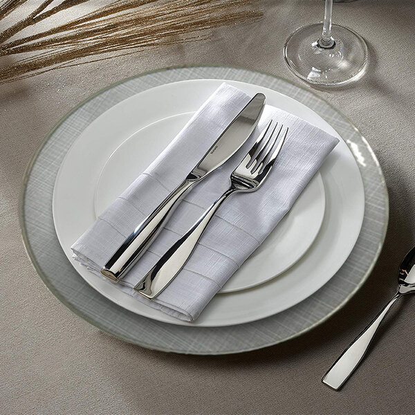 A white Charge It by Jay Laural glass charger plate with silverware and a napkin on it.