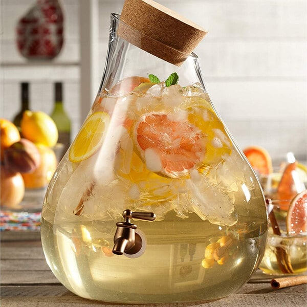 An American Atelier glass teardrop beverage dispenser with a citrus drink inside and a cork top with a tap.