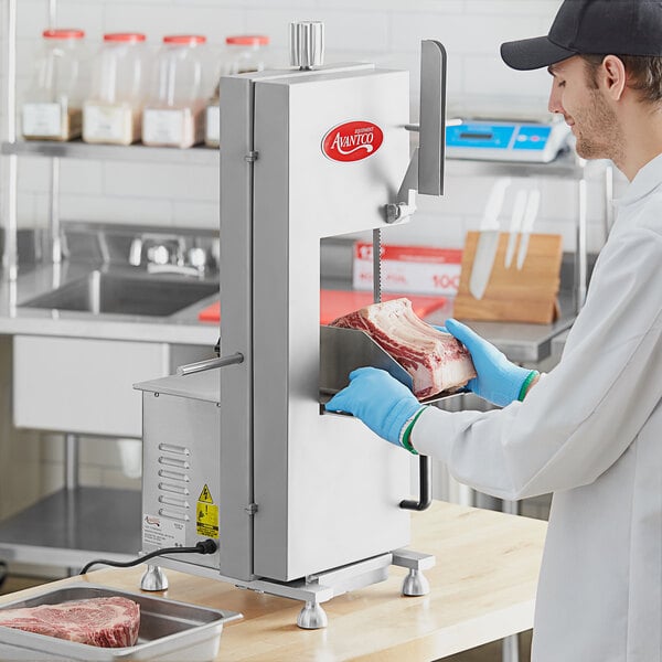 A man in a white coat and blue gloves using an Avantco stainless steel countertop vertical band meat saw to cut meat.