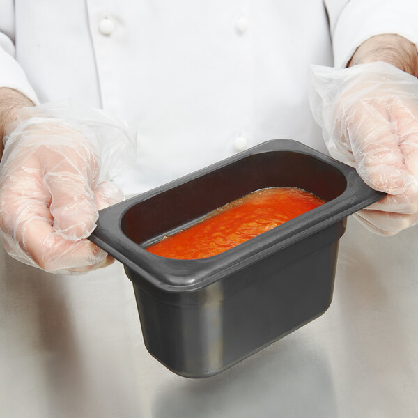 A person in gloves holding a Cambro 1/9 size black plastic food pan filled with red sauce.