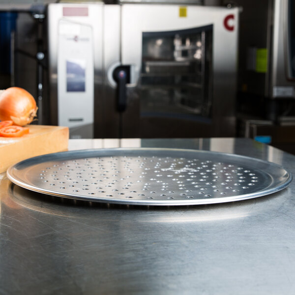 An American Metalcraft heavy weight aluminum pizza pan with holes in it on a cutting board with a tomato and onion slice.