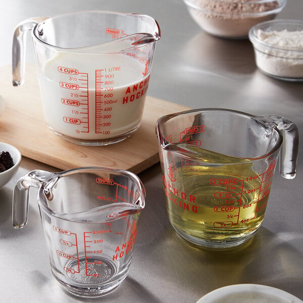 A clear Anchor Hocking measuring cup with red lettering filled with yellow liquid on a counter with a bowl of white powder.