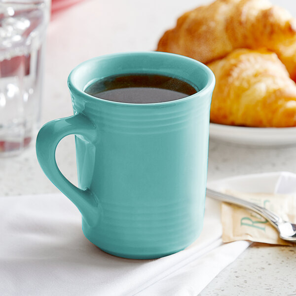 A blue Tuxton Concentrix gala mug filled with coffee next to a croissant.