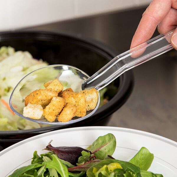 A hand holding a Thunder Group clear polycarbonate salad bar spoon full of croutons over a bowl of lettuce.