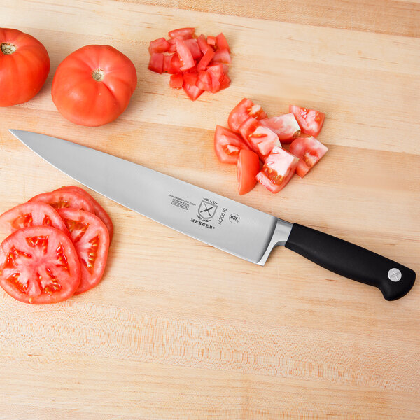 A Mercer Culinary Genesis chef knife cutting a tomato on a cutting board with more tomatoes.