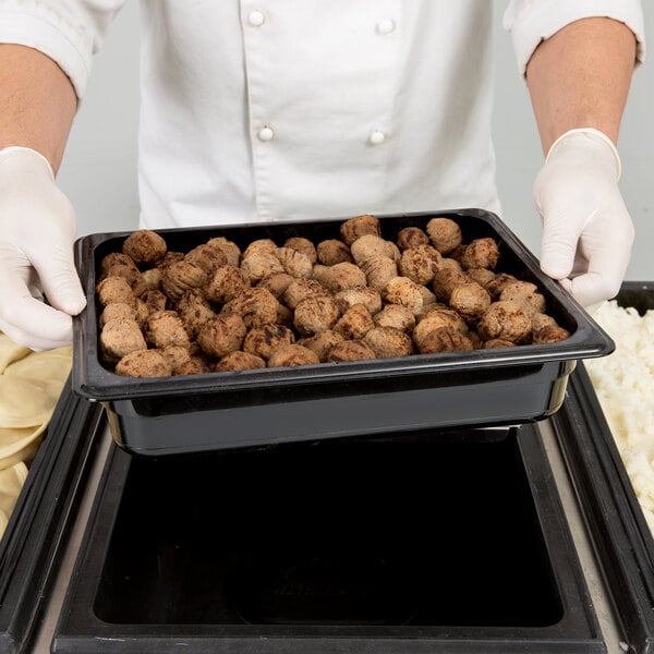 A person in white gloves holding a Cambro black plastic food pan full of meatballs.