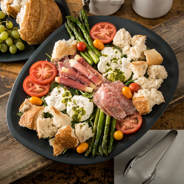 A Carlisle slate melamine platter with asparagus, tomatoes, and bread on a table.