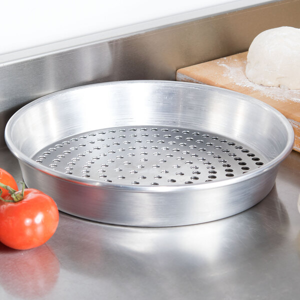 An American Metalcraft heavy weight aluminum pizza pan with perforations next to tomatoes and dough.
