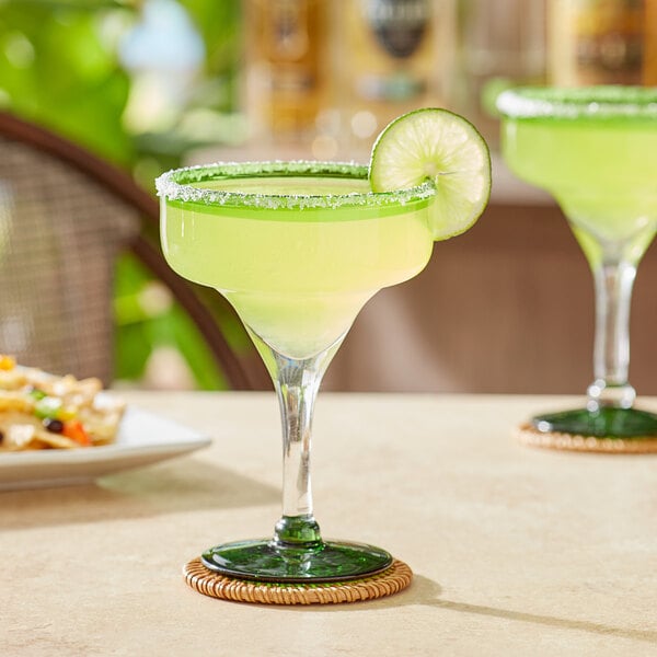Two Acopa margarita glasses with green rims and bases filled with yellow drinks on a table with food.