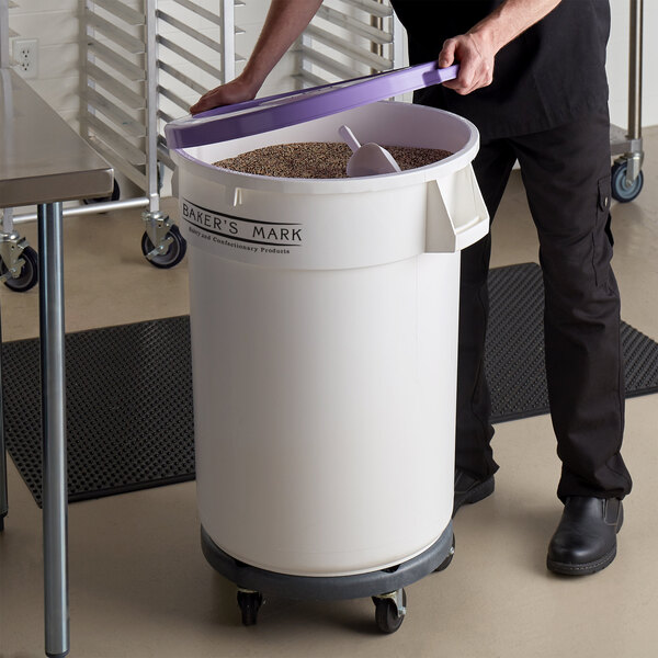 A person using a gray dolly to move a large white container with a purple lid and purple handle.