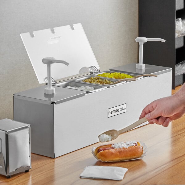 A person using a Nemco stainless steel condiment bar with pumps to prepare a hot dog.