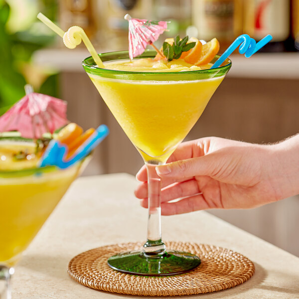 A hand holding an Acopa martini glass with a yellow tropical drink, garnished with a slice of orange and a blue straw.