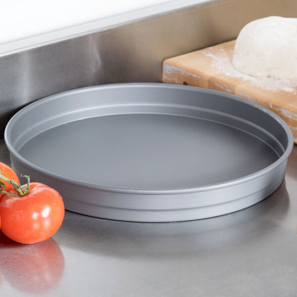 An American Metalcraft hard coat anodized aluminum cake pan on a counter next to pizza dough and tomatoes.