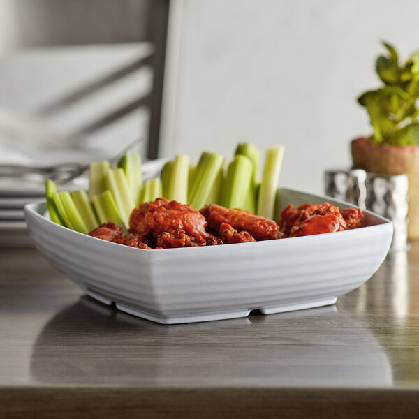 A white Milano square bowl filled with chicken wings and celery on a table.