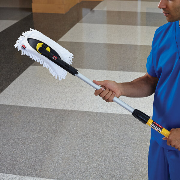A man in blue scrubs holding a Rubbermaid HYGEN white microfiber mop pad on a yellow and black handle.