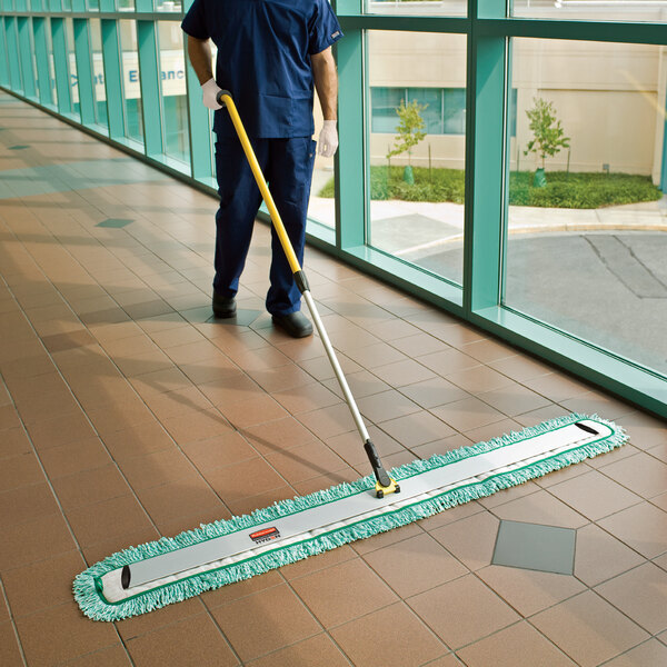 A man in a blue uniform using a Rubbermaid green microfiber fringed dust mop to clean the floor.