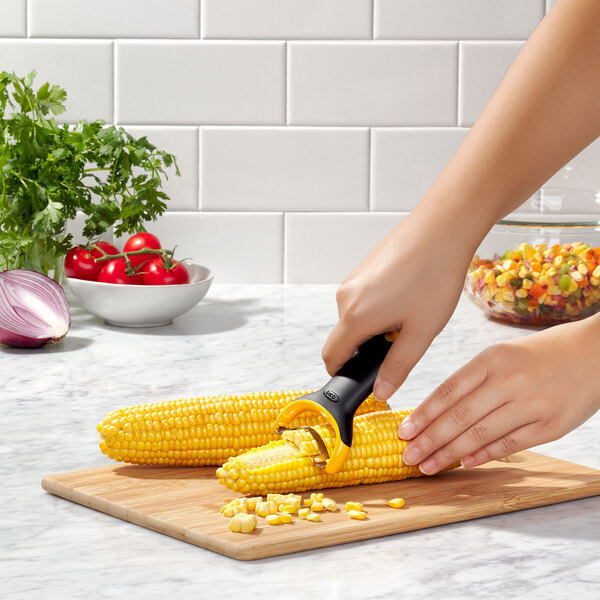 A hand using an OXO "Y" corn peeler to strip corn on a cutting board.
