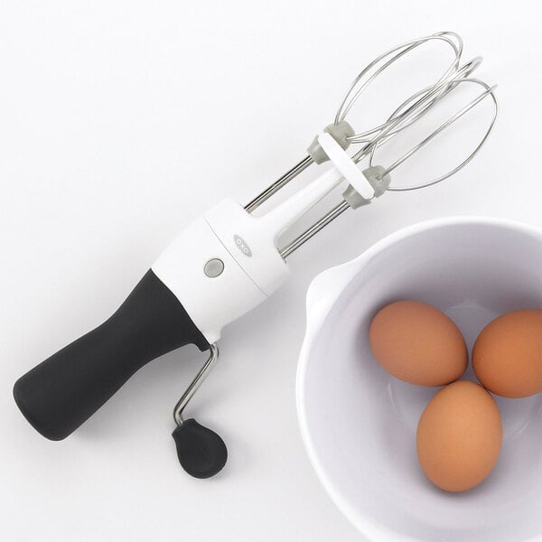 A person using an OXO stainless steel manual crank egg beater to mix eggs in a bowl.