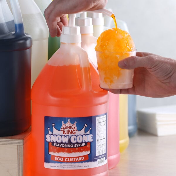 A person using a plastic bottle pump to pour orange liquid into a cup of snow cone.