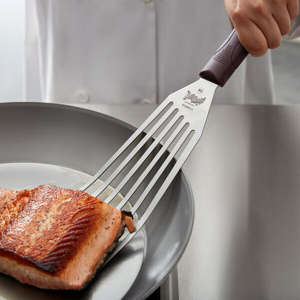 A left-handed Mercer Culinary Hell's Handle slotted turner lifting cooked salmon from a pan.