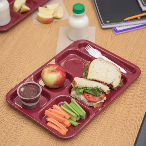 A Carlisle dark cranberry 6 compartment tray on a table with a sandwich, apple, carrots, celery, and a cup of chocolate pudding.