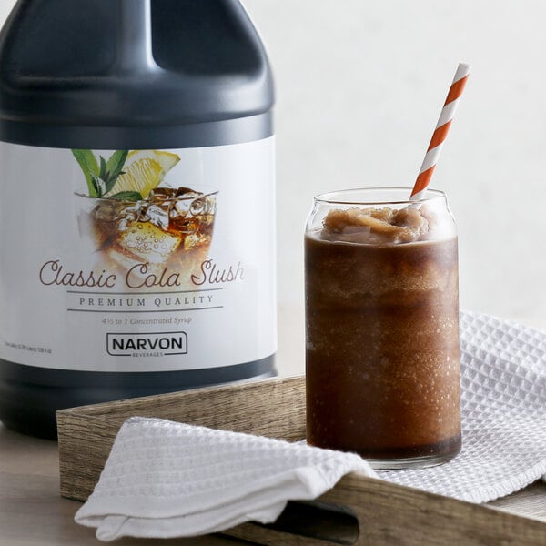A bottle of Narvon Cola Slushy concentrate next to a glass of brown liquid with a straw.