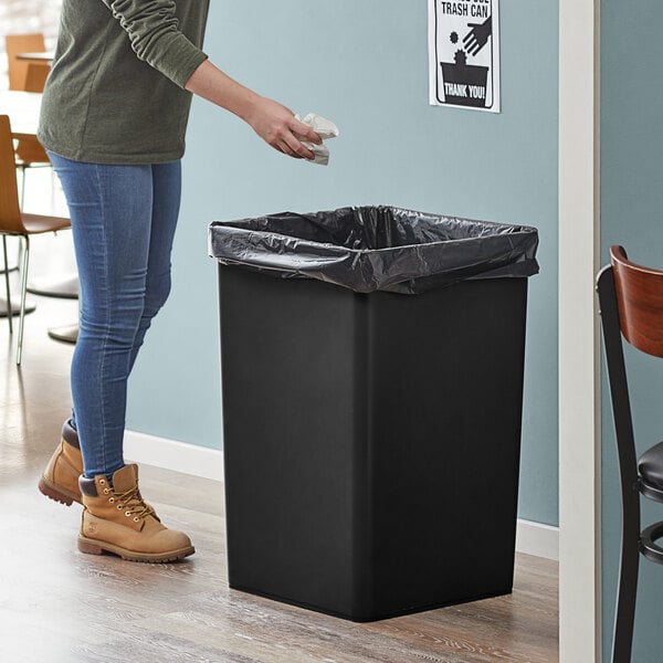 A woman standing next to a Lavex black square trash can in a corporate office cafeteria.