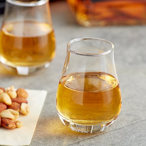 Two Acopa tulip glasses of whiskey on a table with a plate of nuts.