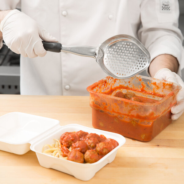 A person using a Vollrath black metal strainer spoon to remove food from a white container.