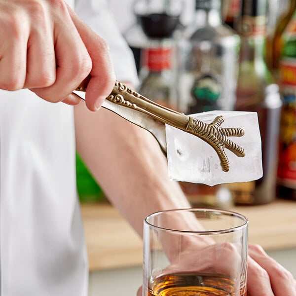 A person using Barfly talon ice tongs to add ice to a glass of amber liquid.