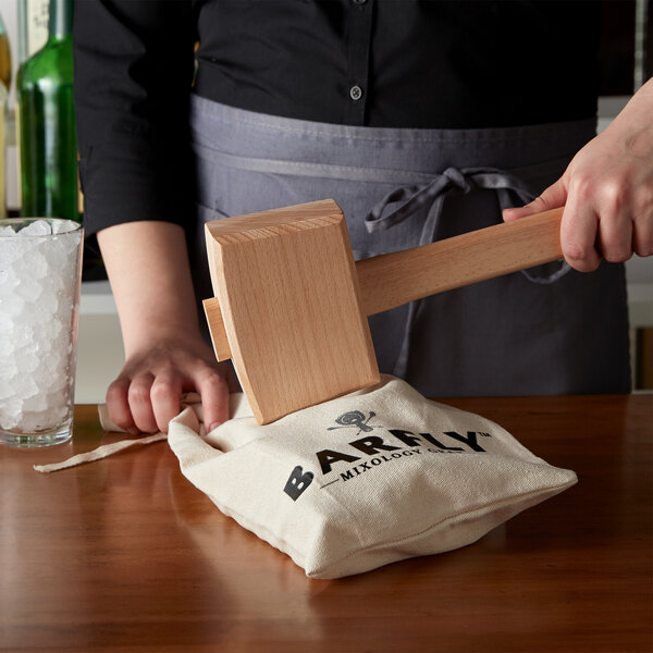 A person holding a Barfly wooden ice mallet over a Lewis canvas bag.