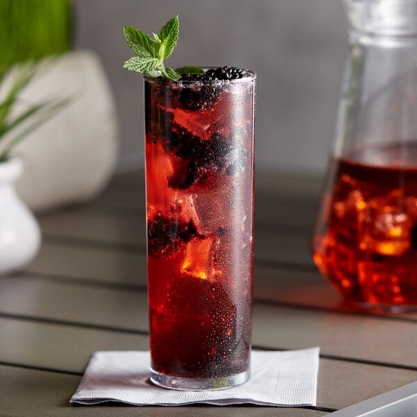 A glass of red liquid with ice and blackberries and mint leaves with a bottle of DaVinci Gourmet Classic Blackberry Fruit Syrup on a table.