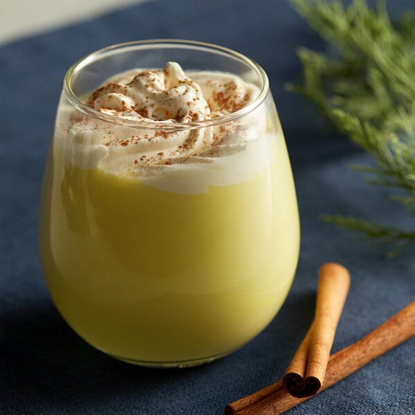 A glass of yellow liquid with whipped cream and cinnamon next to DaVinci Gourmet Eggnog Syrup and cinnamon sticks.