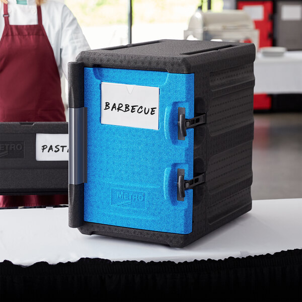 A black Metro Mightylite food pan carrier with a blue door on a table in an outdoor catering setup.