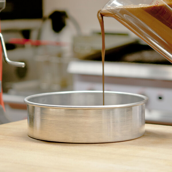 A person pouring liquid into an American Metalcraft aluminum round cake pan on a professional kitchen counter.