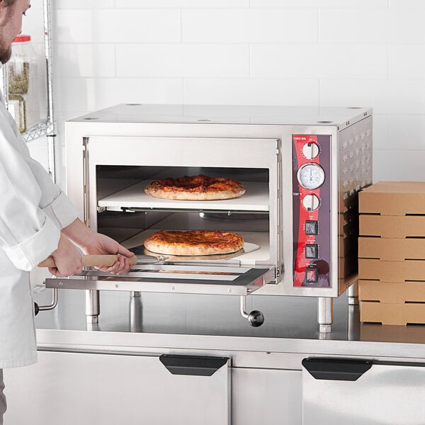 A man in a white coat putting pizzas into an Avantco double deck countertop pizza oven.