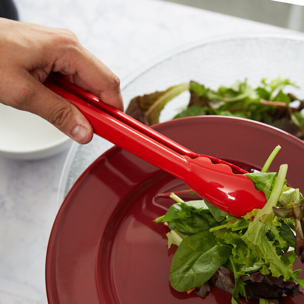 A hand holding a Carlisle red plastic utility tong over a salad