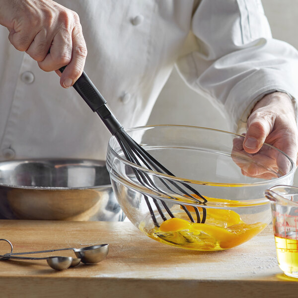 A hand using a Thunder Group black nylon piano whisk to mix eggs in a glass bowl.