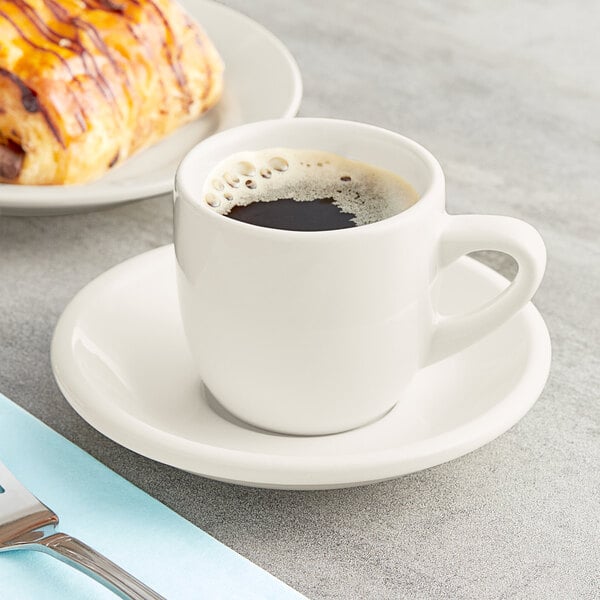 An Acopa ivory espresso cup filled with brown liquid on a saucer with a pastry.