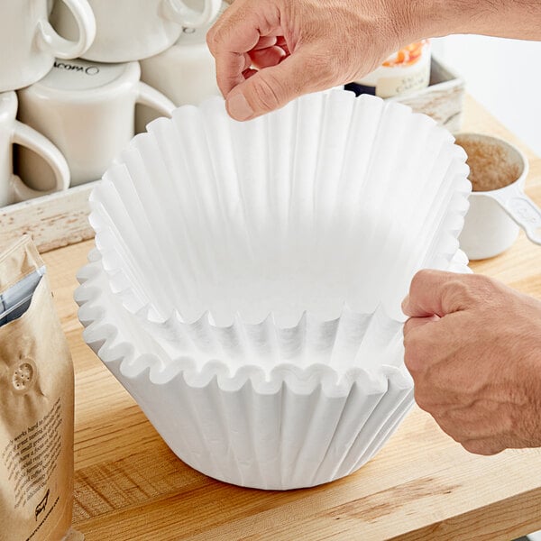 A person holding a Fetco coffee filter over a stack of coffee cups.