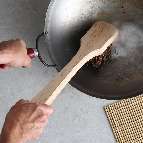 A hand using a Thunder Group Palmyra bristled wok brush with a wood handle to clean a pan.