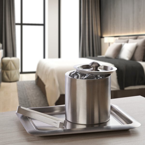 A silver stainless steel ice bucket with ice in it and tongs on a tray.
