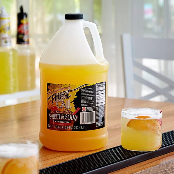 A jug of Finest Call sweet and sour concentrate on a yellow table next to a glass of yellow liquid.