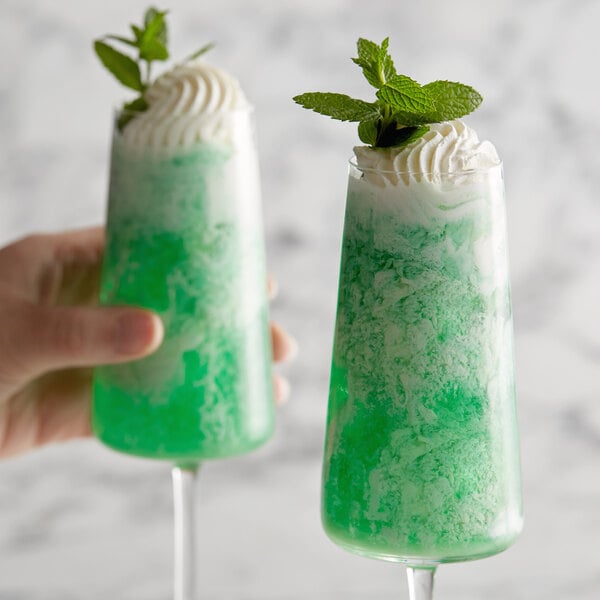 A hand holding two glasses of green and white DaVinci Gourmet Sugar Free Peppermint Paddy drinks with mint leaves.