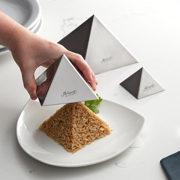 A hand holding a pyramid shaped container over a plate of brown rice.