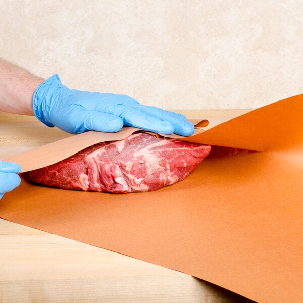 Blue gloved hands wrapping a piece of meat on a Choice pink butcher paper sheet.