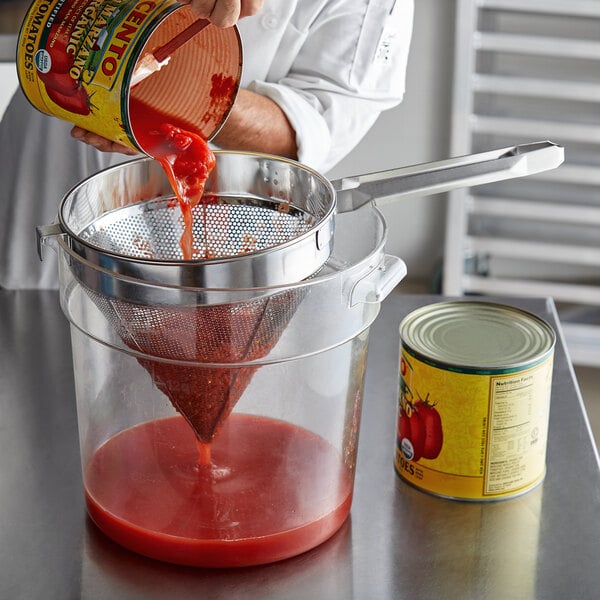 A person pouring tomato sauce into a container using a Choice Coarse China Cap strainer.