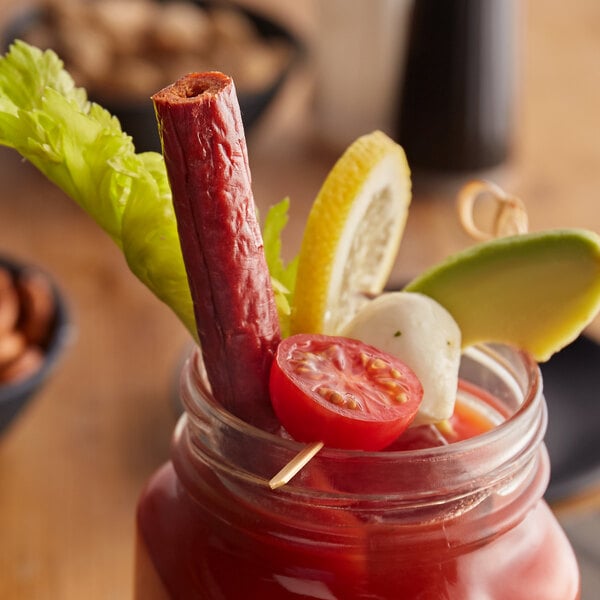 A glass jar with a Bloody Mary with Benny's Original Meat Straws, vegetables, and a tomato on a table.