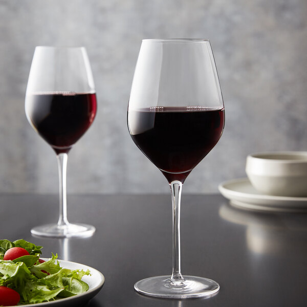 Two Stolzle Bordeaux wine glasses filled with red wine on a table with salad.