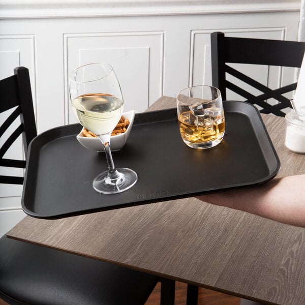 A person holding a Cambro black non-skid serving tray with two glasses of white wine and a bowl of ice cubes.